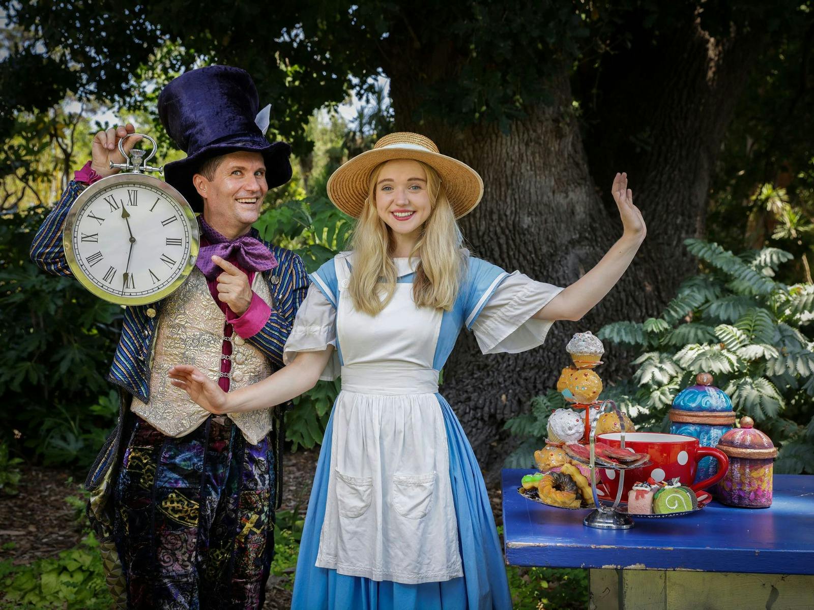 Two people in costume, one with a clock and the other in a blue dress, pose by desserts.