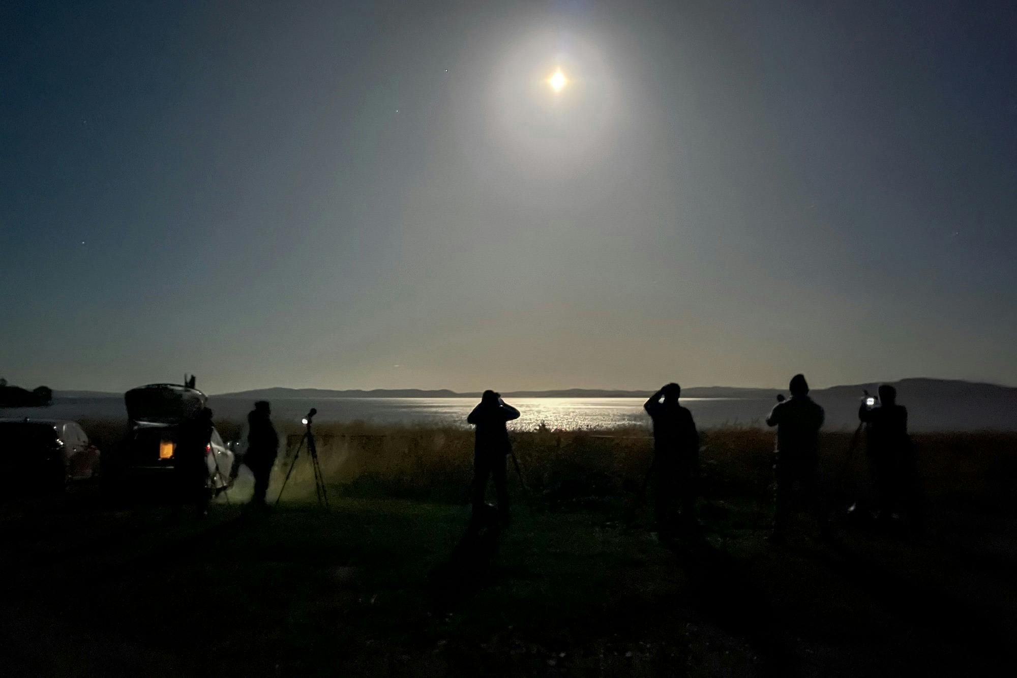 A group of photographers with tripods watches a bright full moon above a dark horizon at night.