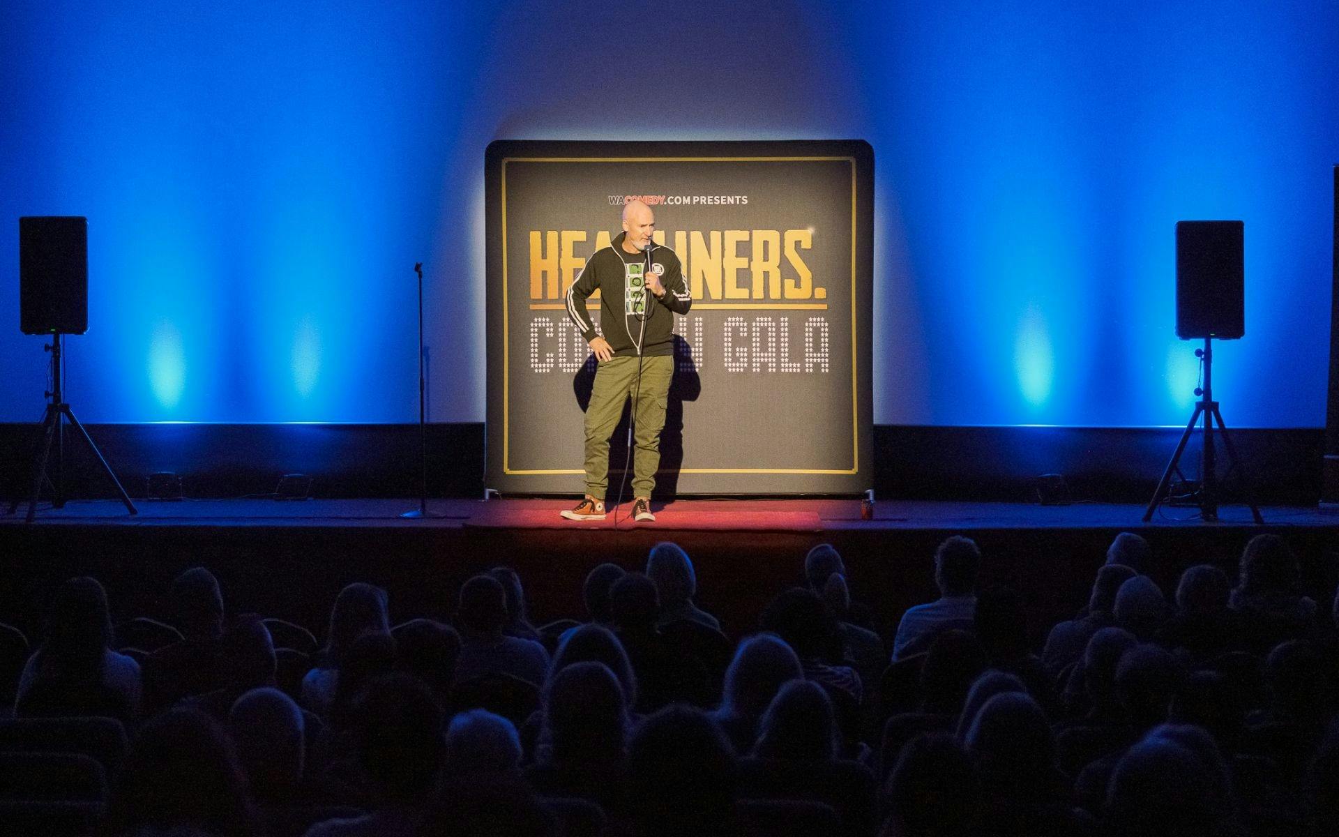 A standup comedian speaks into a microphone onstage, a sign reading "Headliners Comedy Gala" behind.