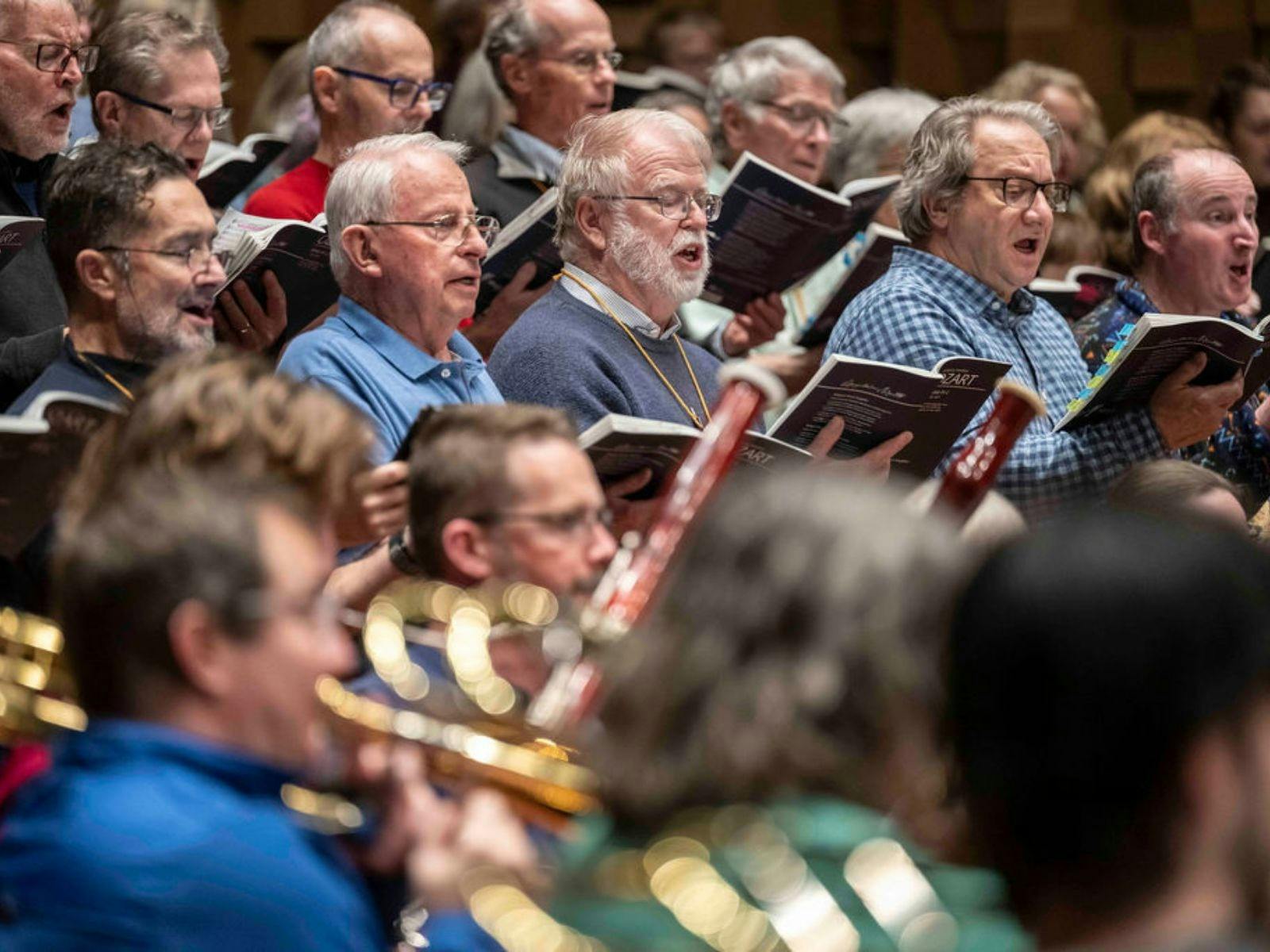 Choir members singing with music sheets and musicians playing brass instruments in foreground.