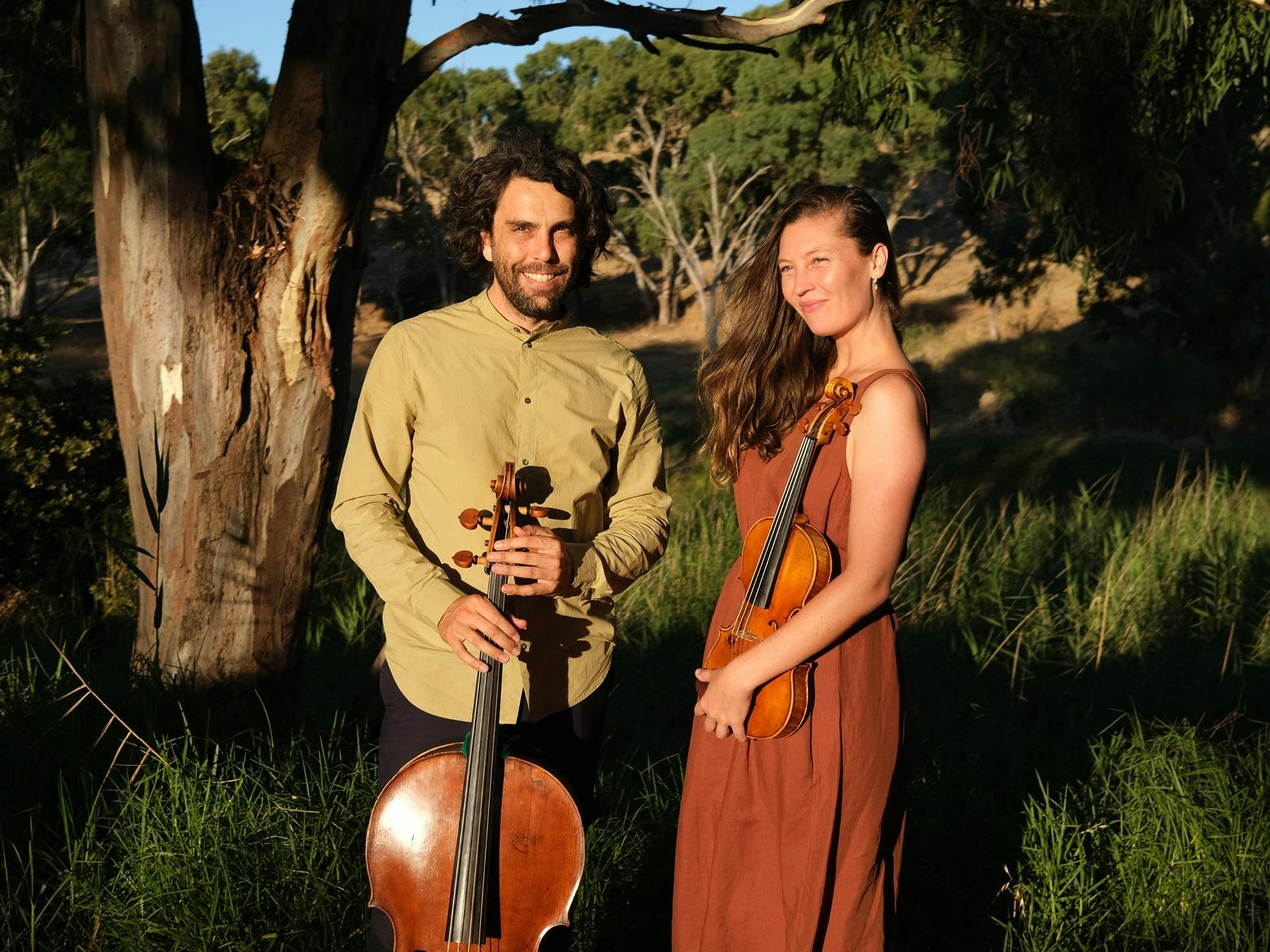 Two people standing outdoors holding a cello and a violin in warm sunlight. Two people standing outdoors holding a cello and a violin in warm sunlight.