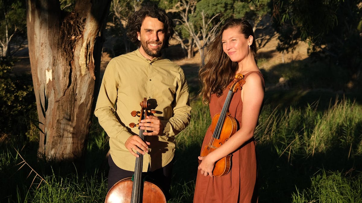 Two people standing outdoors holding a cello and a violin in warm sunlight.