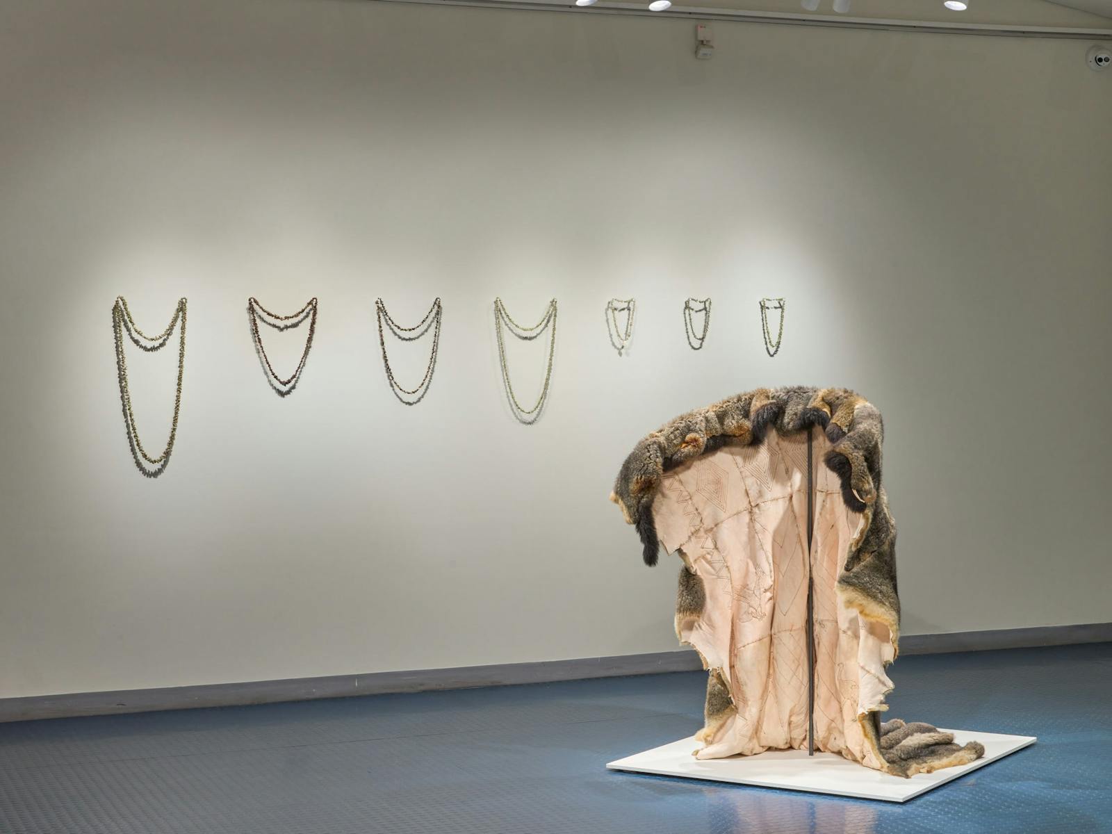In an exhibition, several necklaces hang on the wall and a fur coat is draped in the foreground.