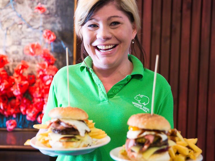 Smiling woman in Wombat Hotel uniform, holding up two plates of burgers and chips.