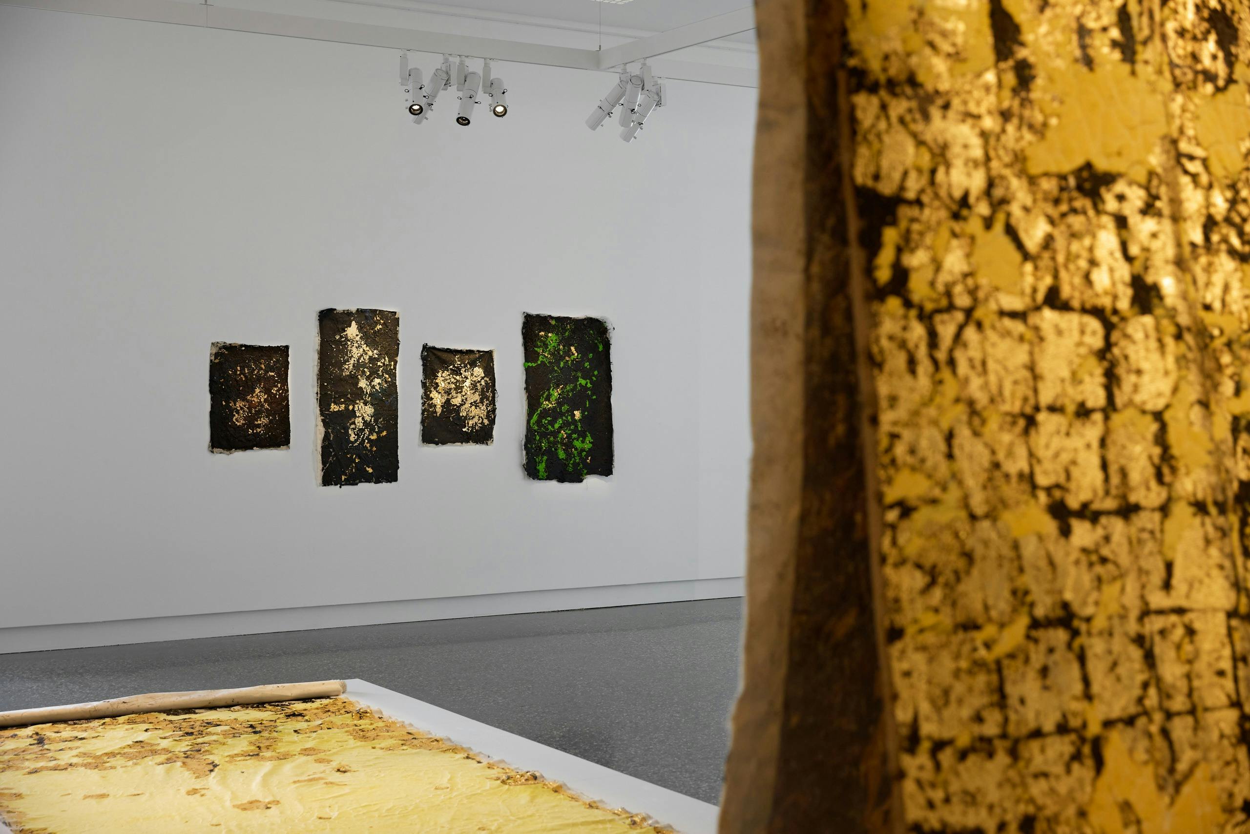 Installation view of four artworks on white wall framed by a sculpture with gold surface.