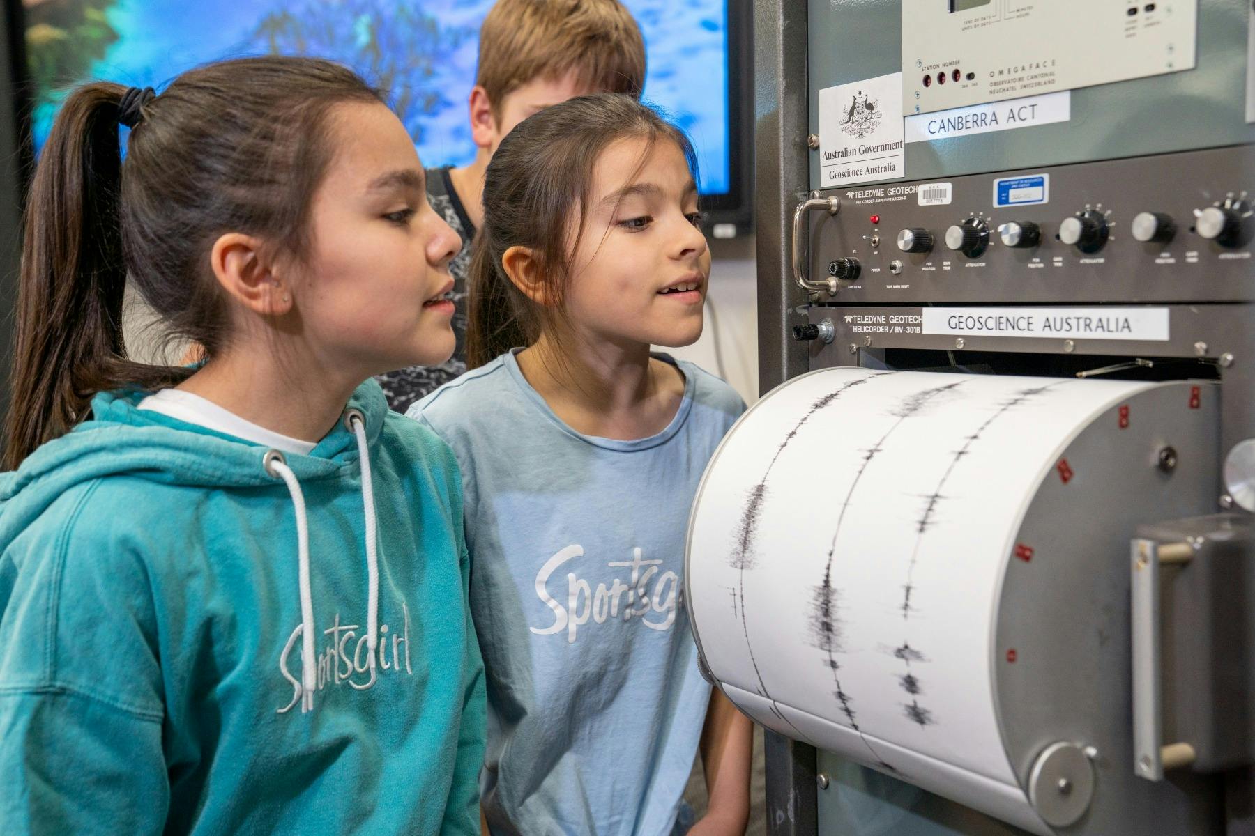 Two pre-teen girls look at a cylindrical drum that has white paper with black wave signals on it.