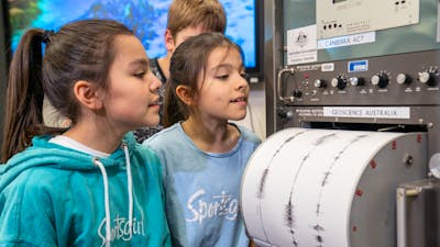 Two pre-teen girls look at a cylindrical drum that has white paper with black wave signals on it.