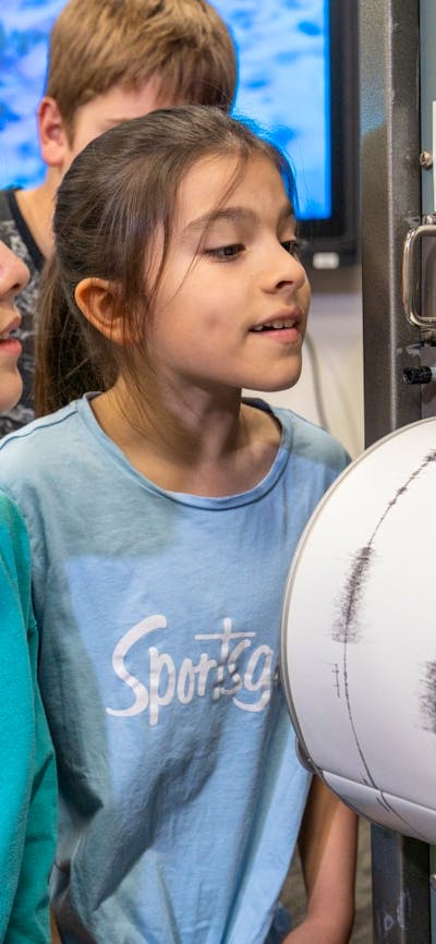 Two pre-teen girls look at a cylindrical drum that has white paper with black wave signals on it.