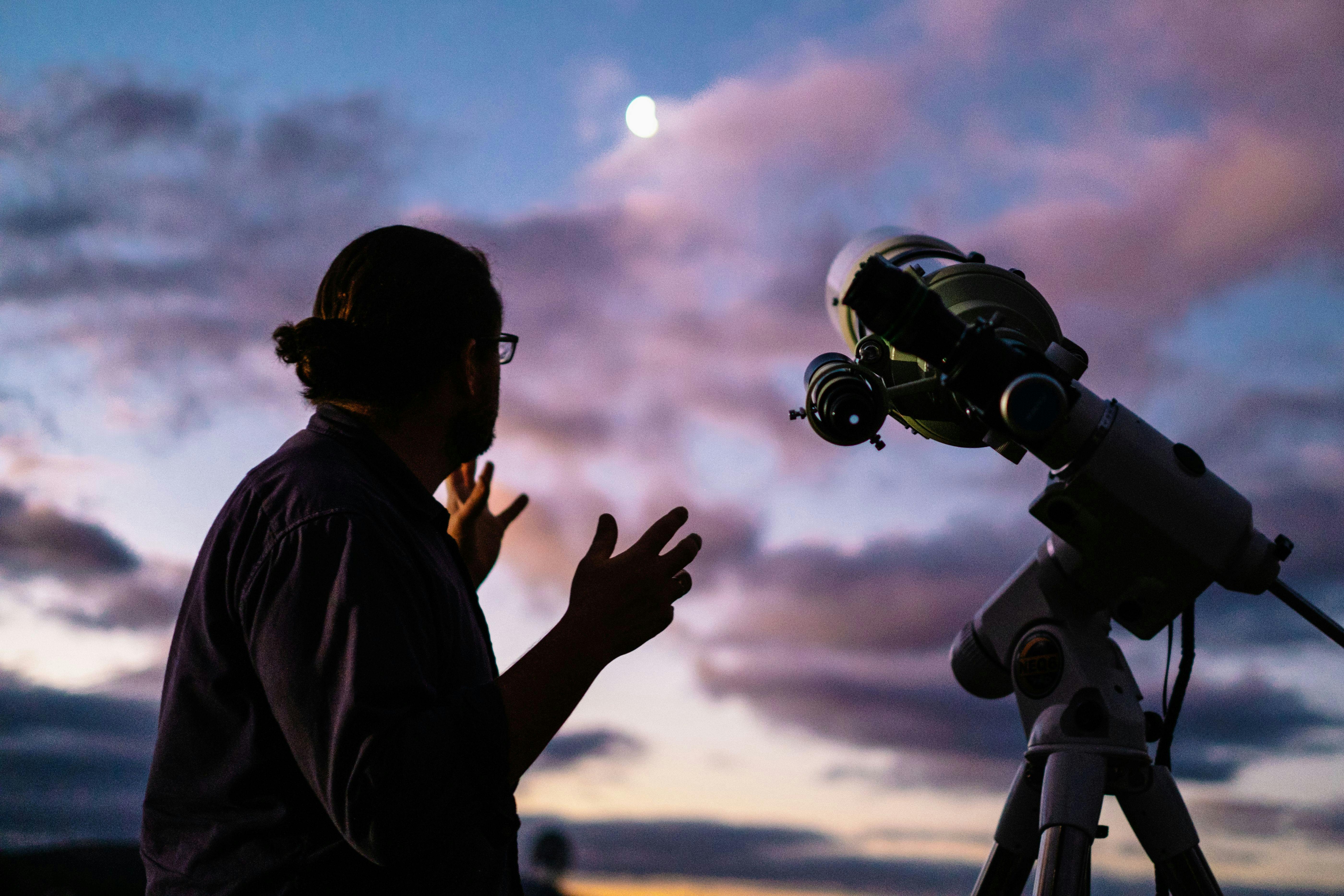 Man looks towards the moon with setting sun next to telescope