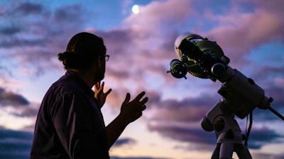 Man looks towards the moon with setting sun next to telescope