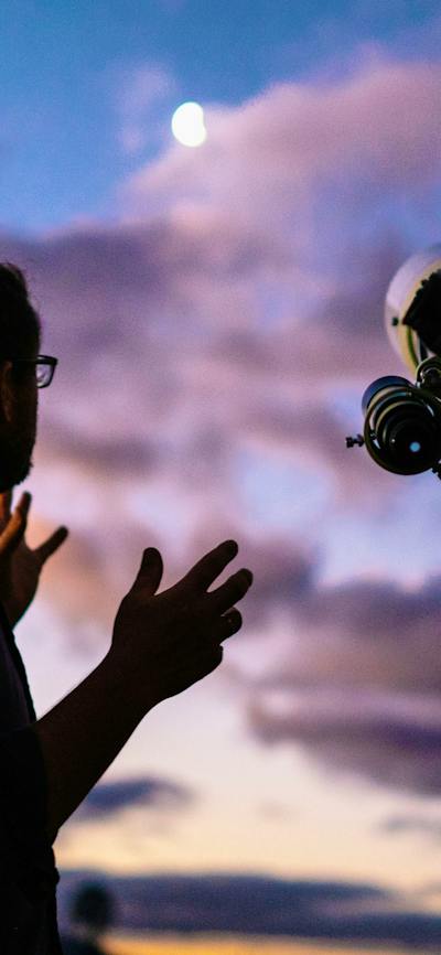 Man looks towards the moon with setting sun next to telescope