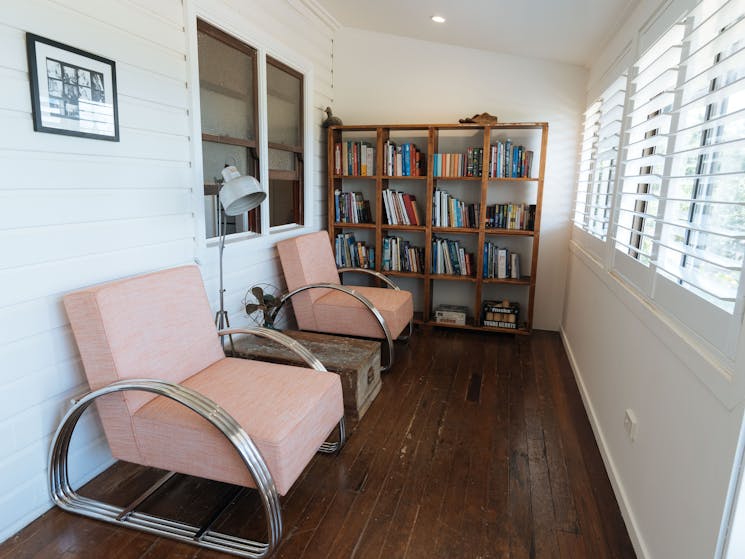A reading room with a bookcase and two pink occasional chairs looking out two windows with blinds