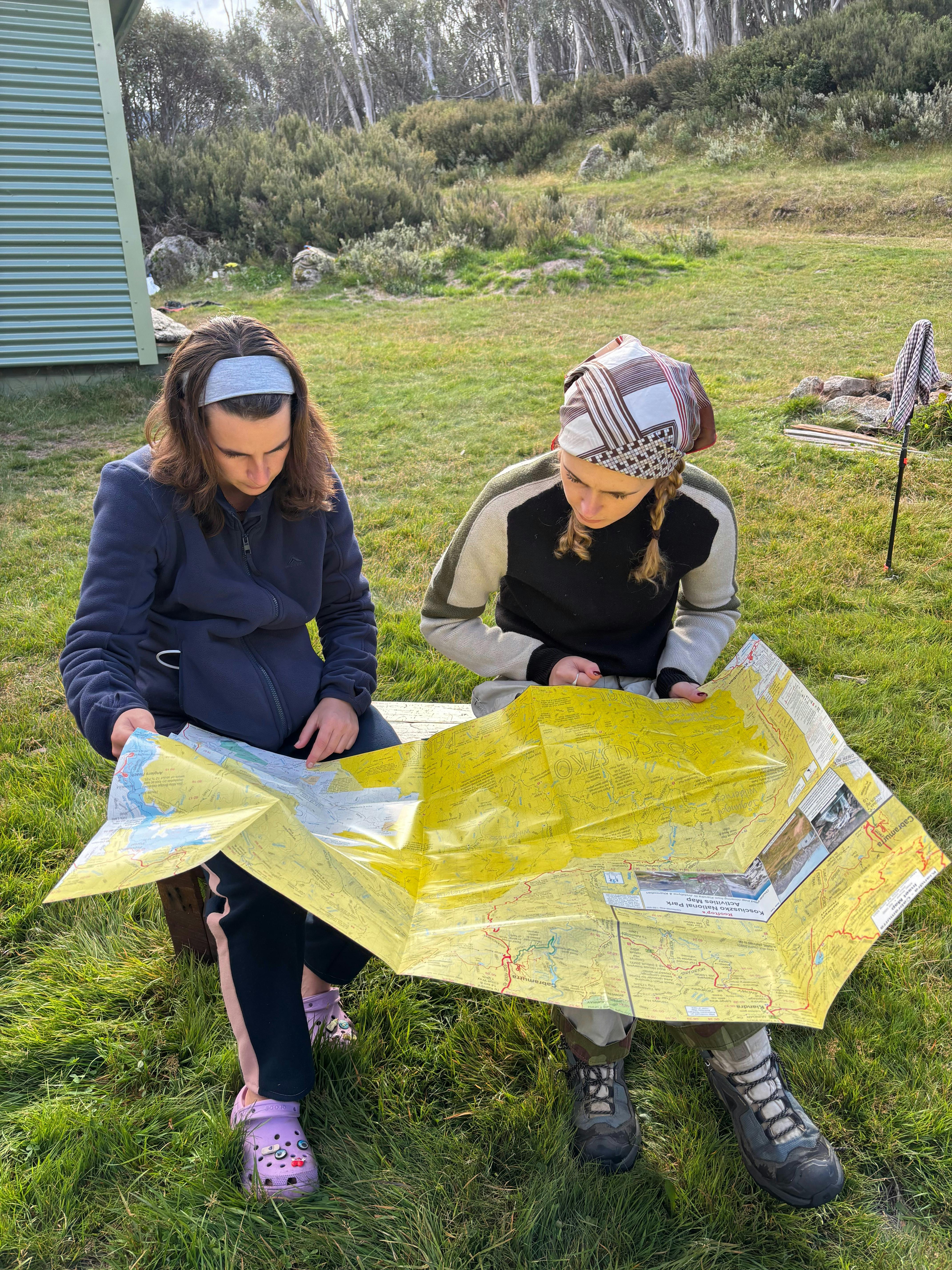 Two women reading a yellow map in front of a small wooden hut.