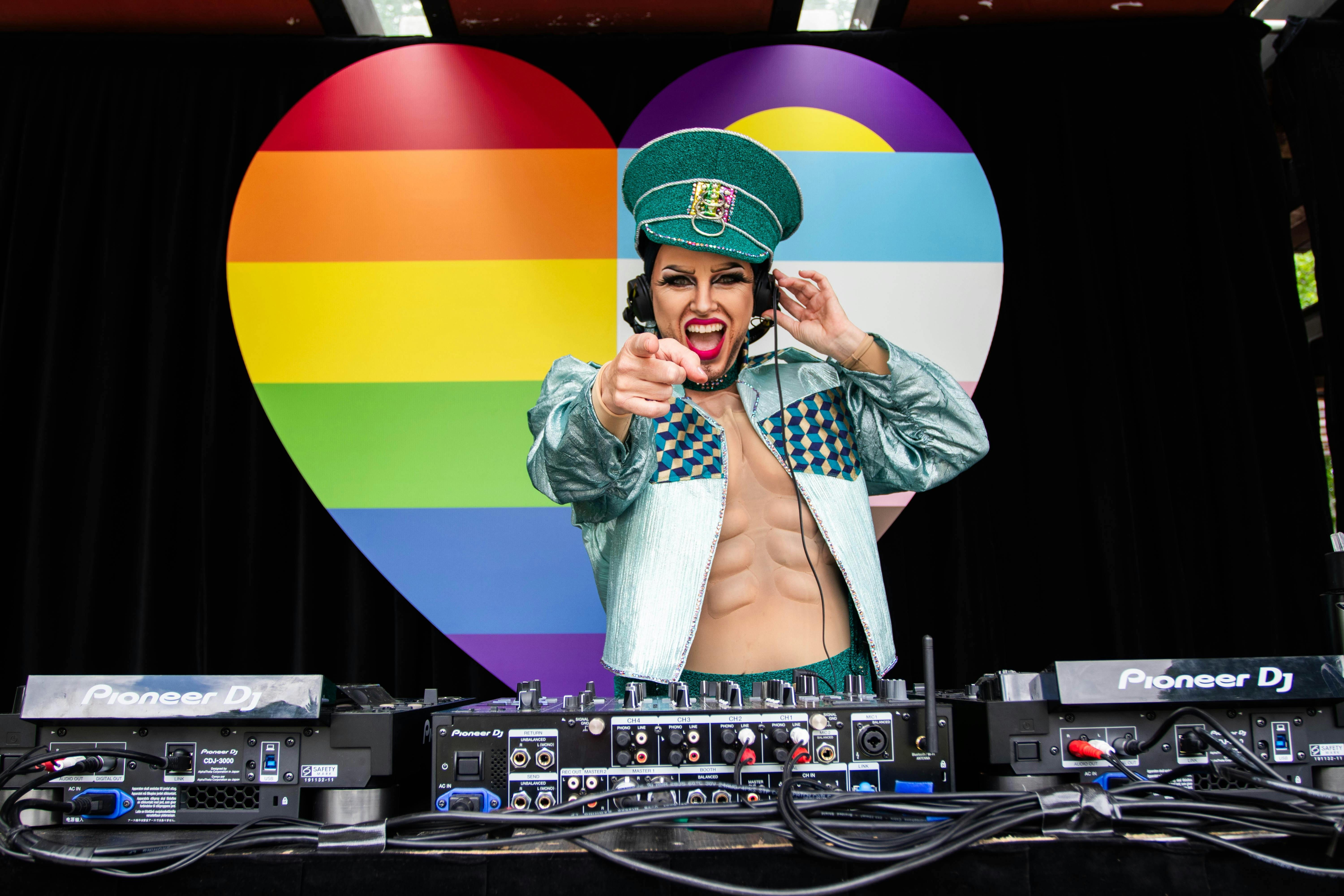 Dj Sexy Galexy boi drag performer and DJ playing music at Fair day Large Rainbow Heart in the backgr