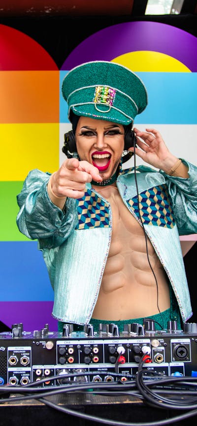 Dj Sexy Galexy boi drag performer and DJ playing music at Fair day Large Rainbow Heart in the backgr