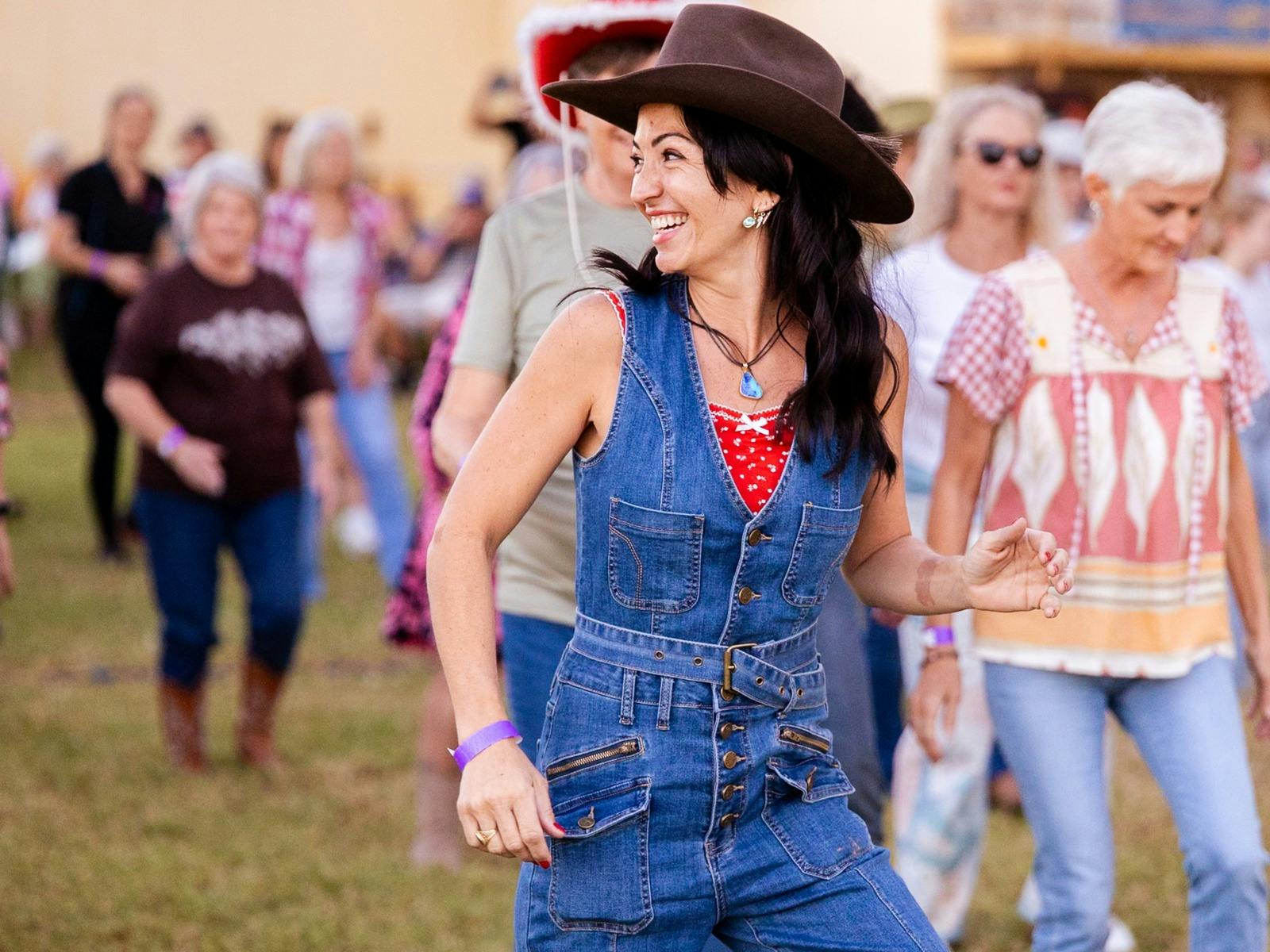 Woman line dancing in the crowd wearing dark blue overalls and brown cowgirl hat