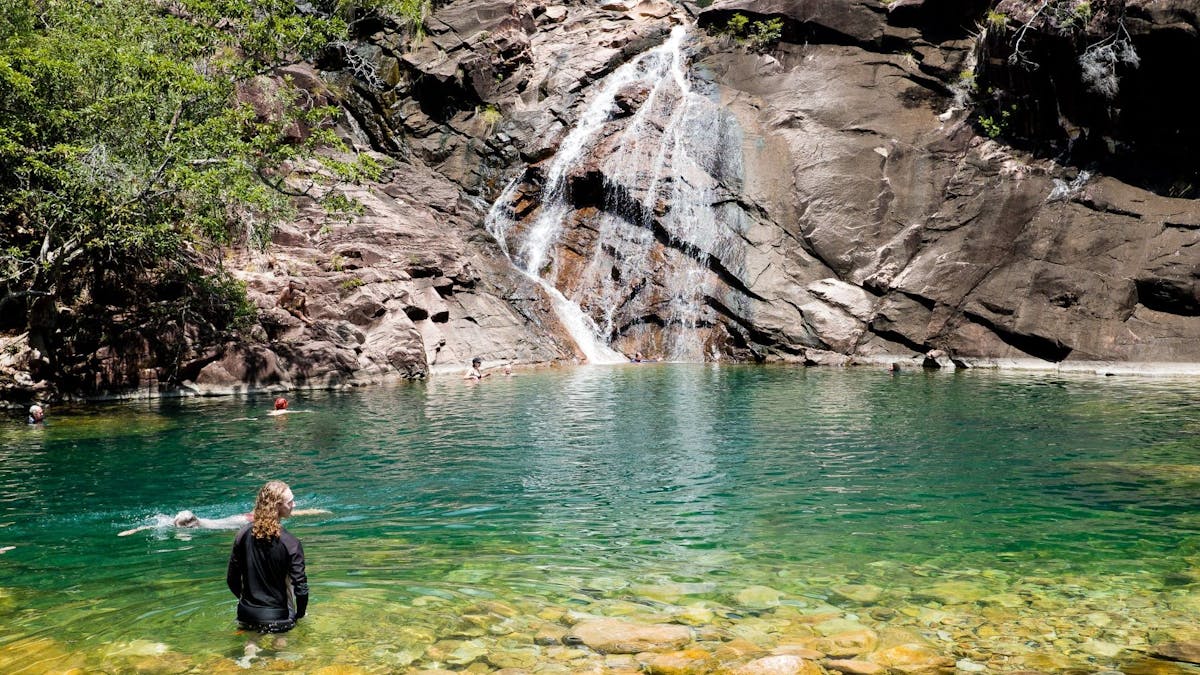 A person swims and stares across stunning aqua coloured water towards the cascading Zoe Falls