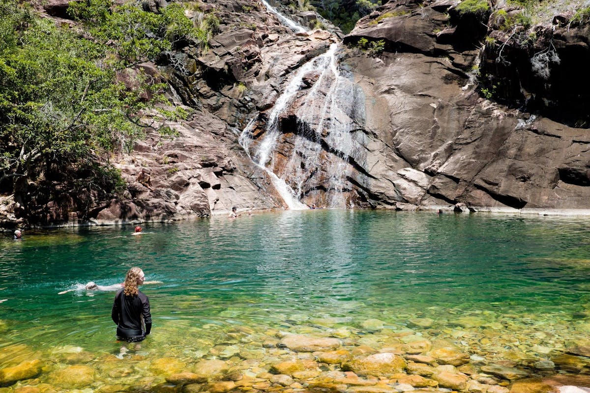 A person swims and stares across stunning aqua coloured water towards the cascading Zoe Falls