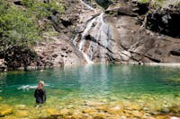 A person swims and stares across stunning aqua coloured water towards the cascading Zoe Falls