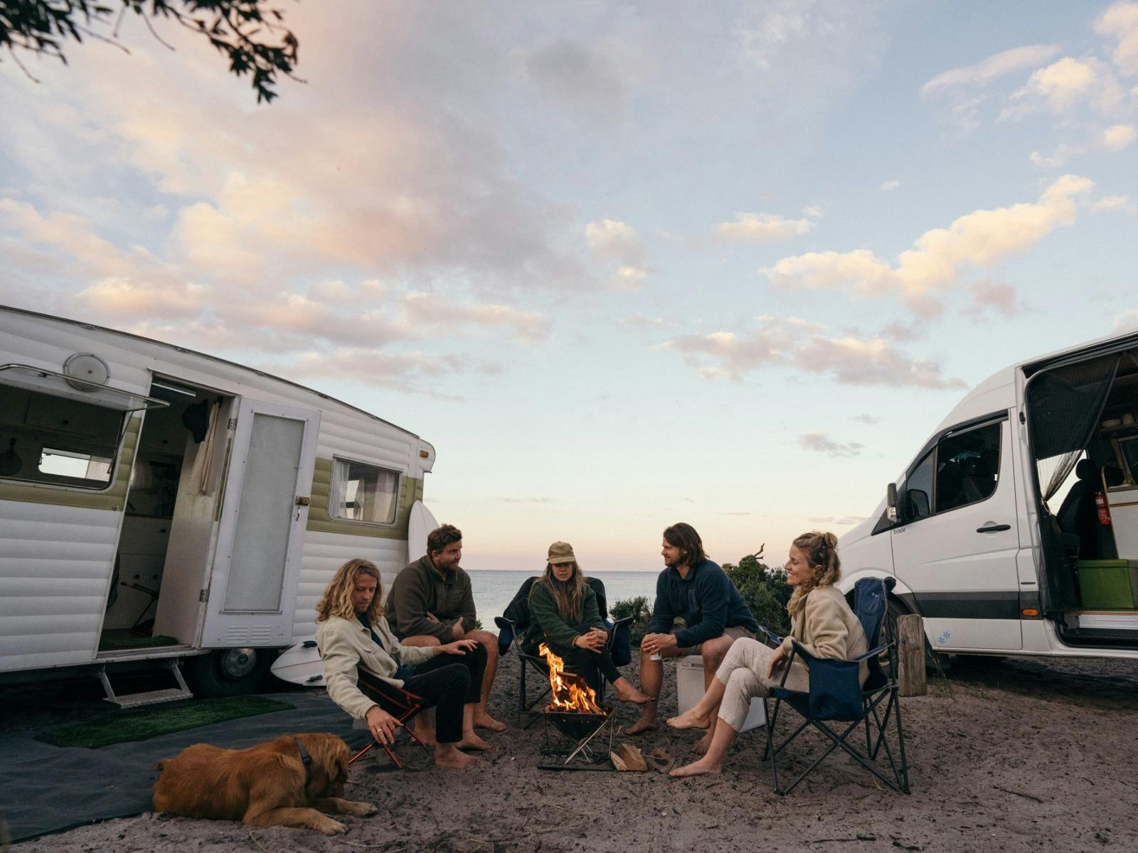 A group of friends and their dog sit in front of a campfire at the beach surrounded by their 2 RVs