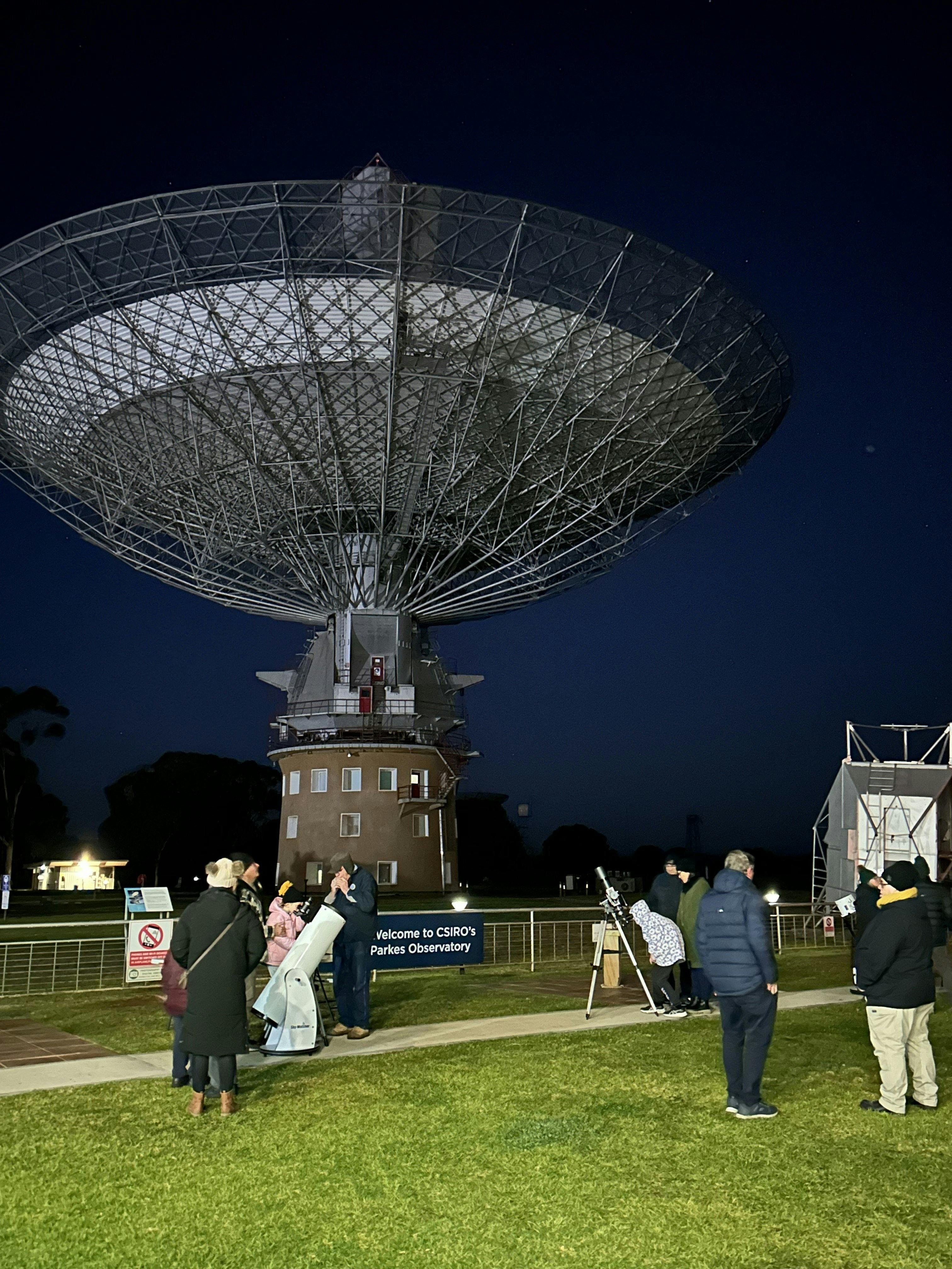 Night view of people looking through optical telescopes with Murriyang in the background