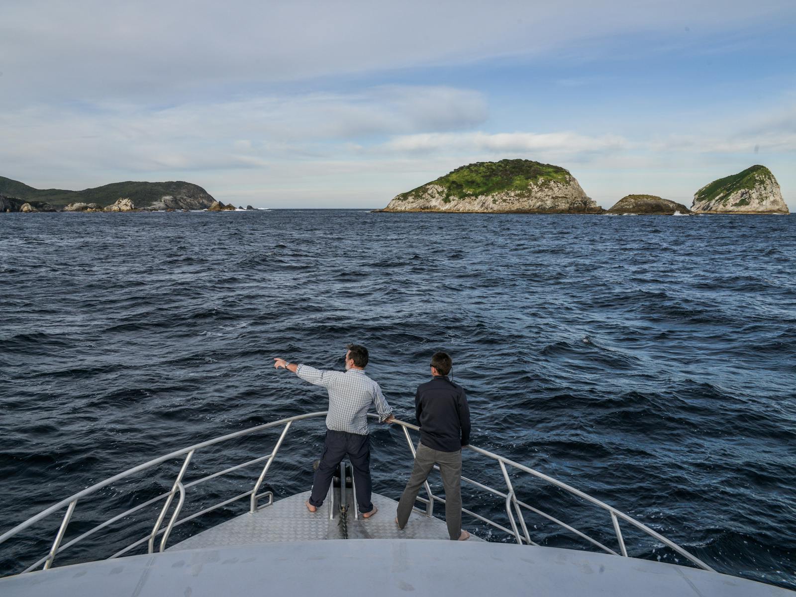 Two people on the bow of a boat, one is pointing and they are looking at islands in the distance.