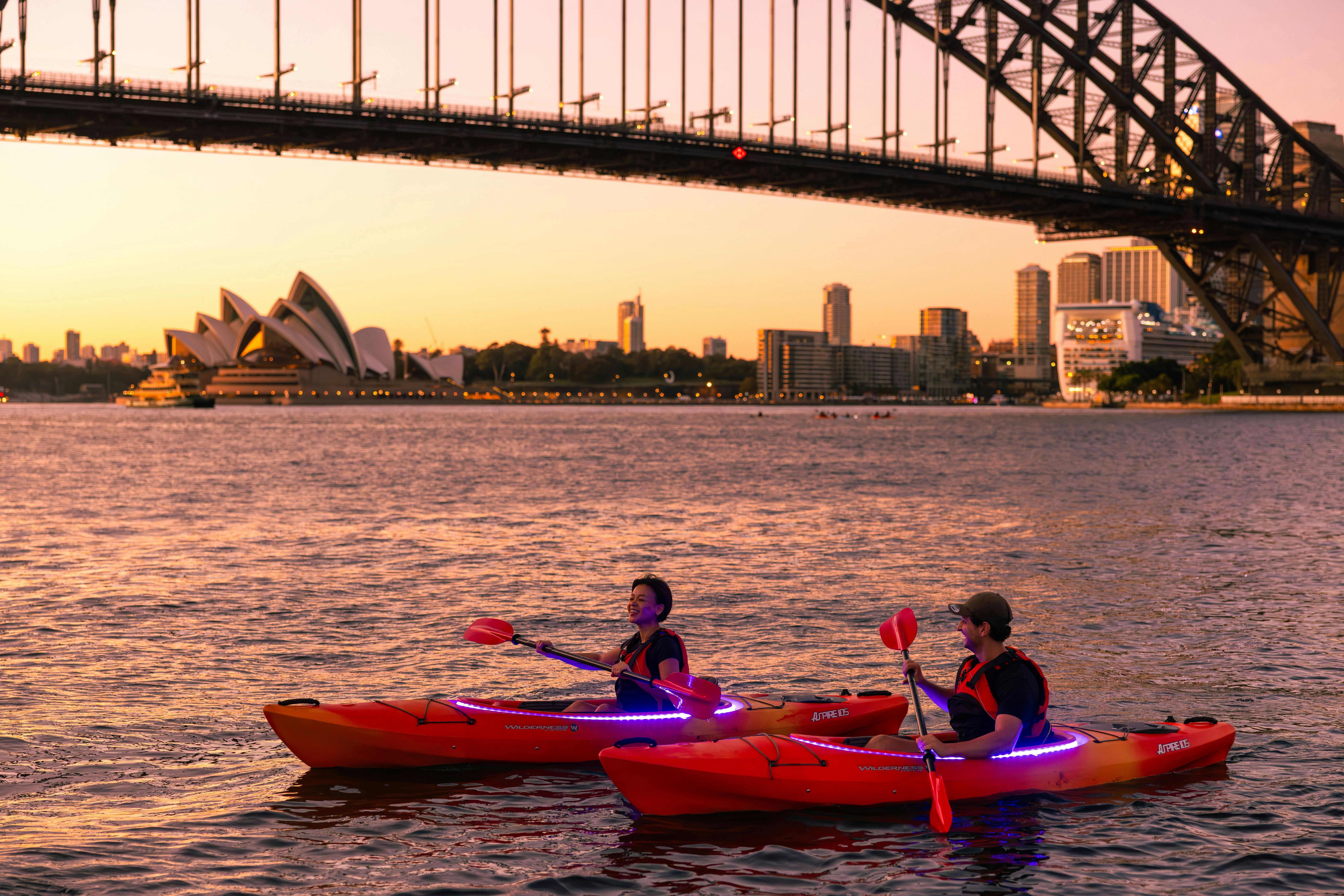 Two people paddling at sunrise with the Sydney Harbour bridge and Opera house with a golden glow