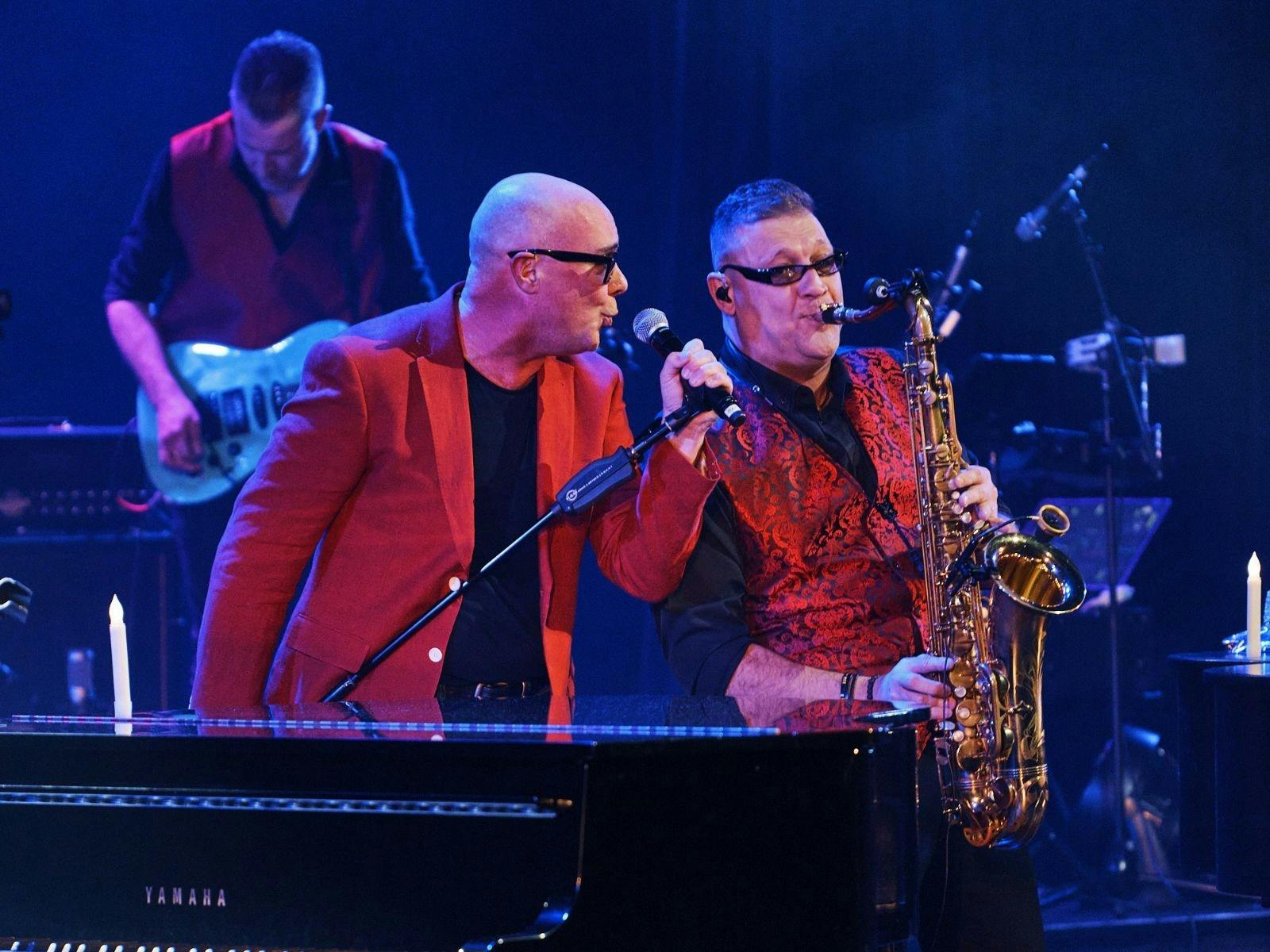 2 men in red jackets 1 with microphone other with saxophone singing 2 men in red jackets 1 with microphone other with saxophone singing
