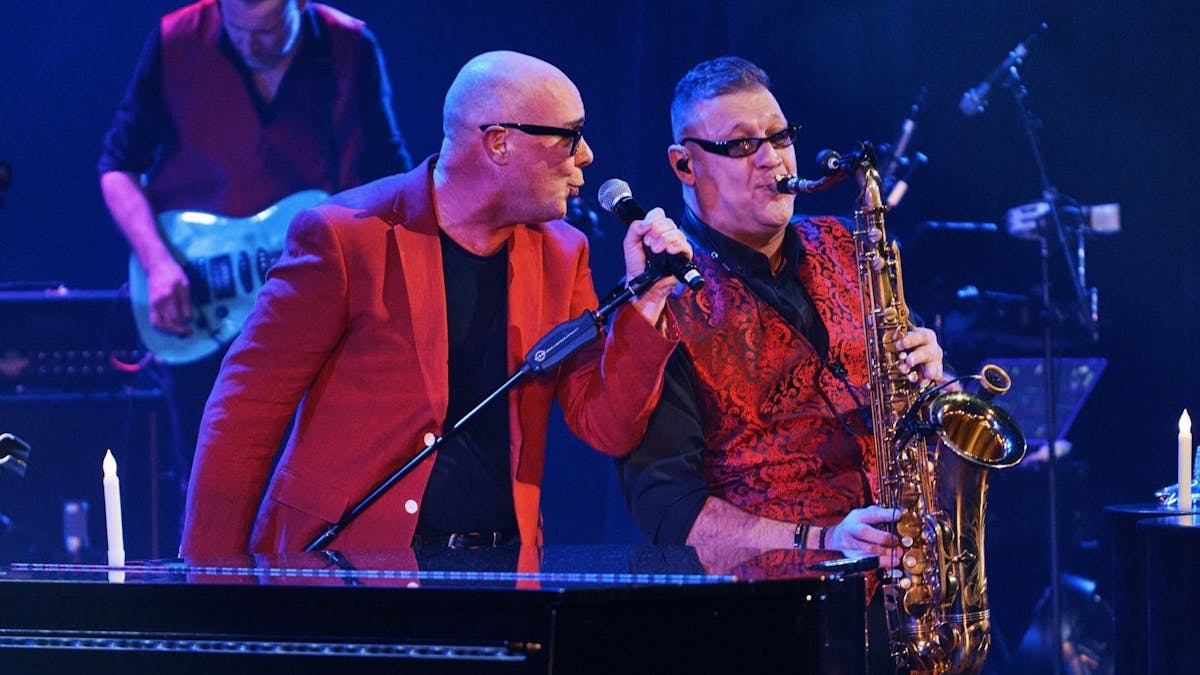 2 men in red jackets 1 with microphone other with saxophone singing