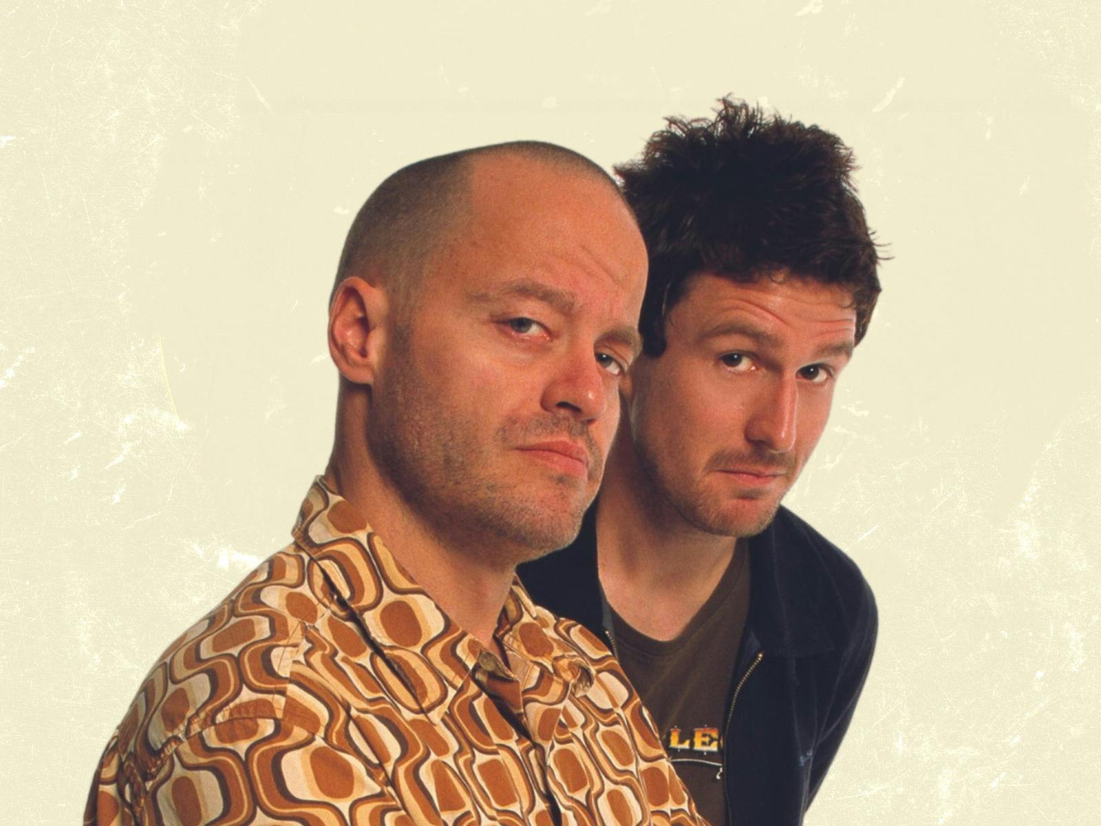 Two men looking at the camera, one wears black the other patterned orange