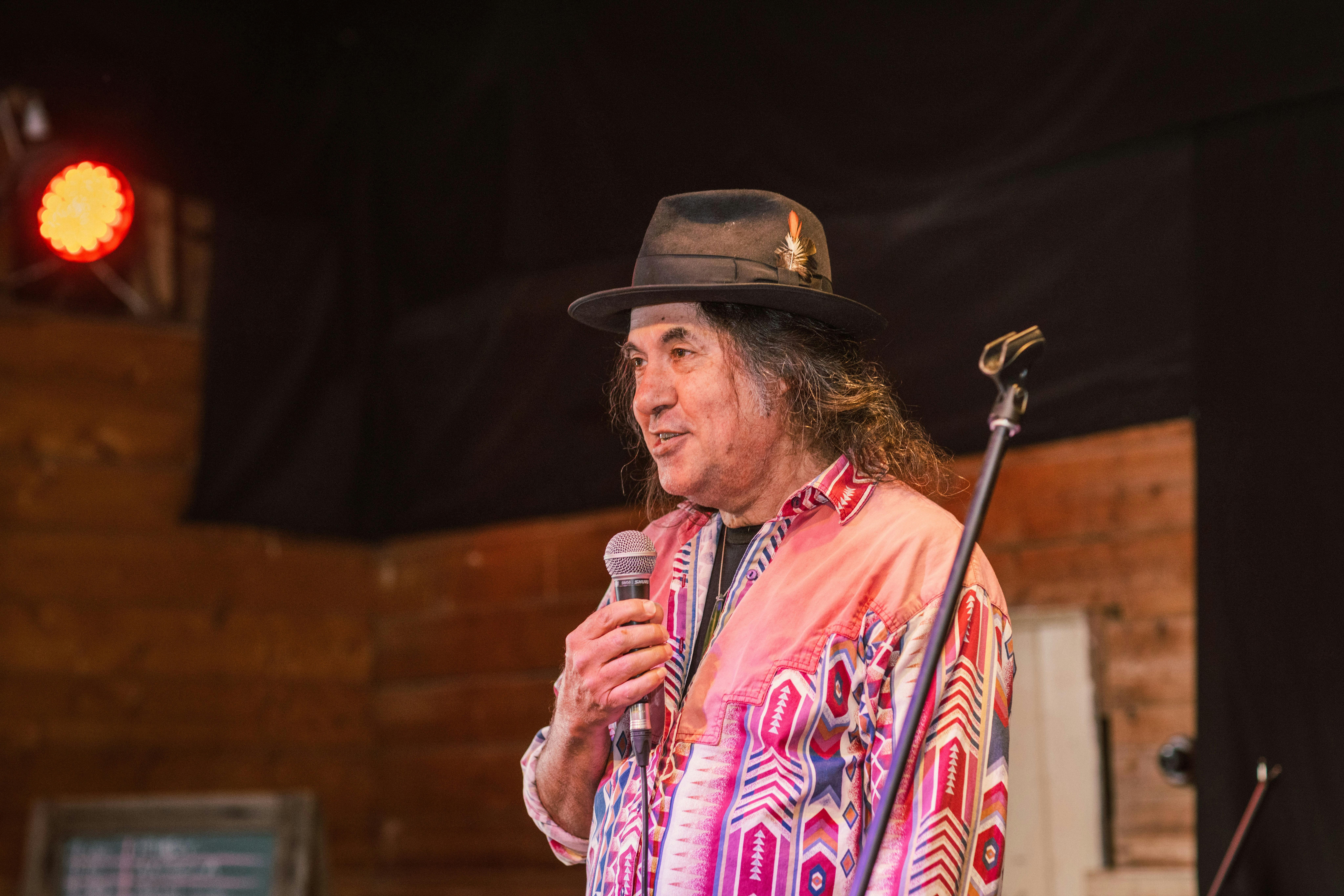 A man in a pink and red shirt is speaking into the mic on stage.