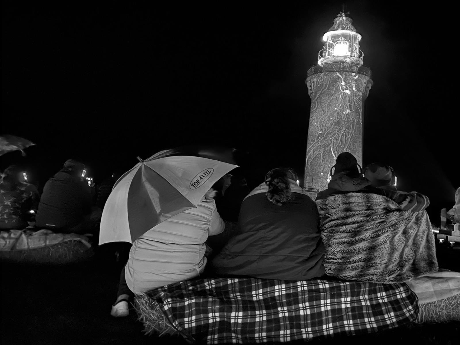 People rugged up with an umbrella watching a projection onto Table Cape lighthouse.