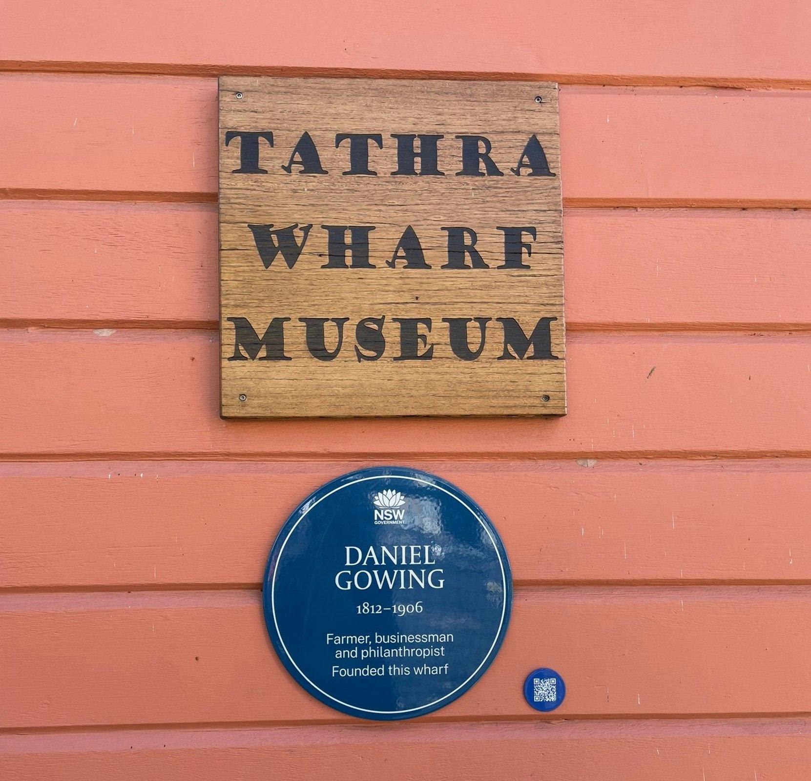 Blue plaque on red wooden horizontal slat wall, under sign saying 'Tathra Wharf Museum'