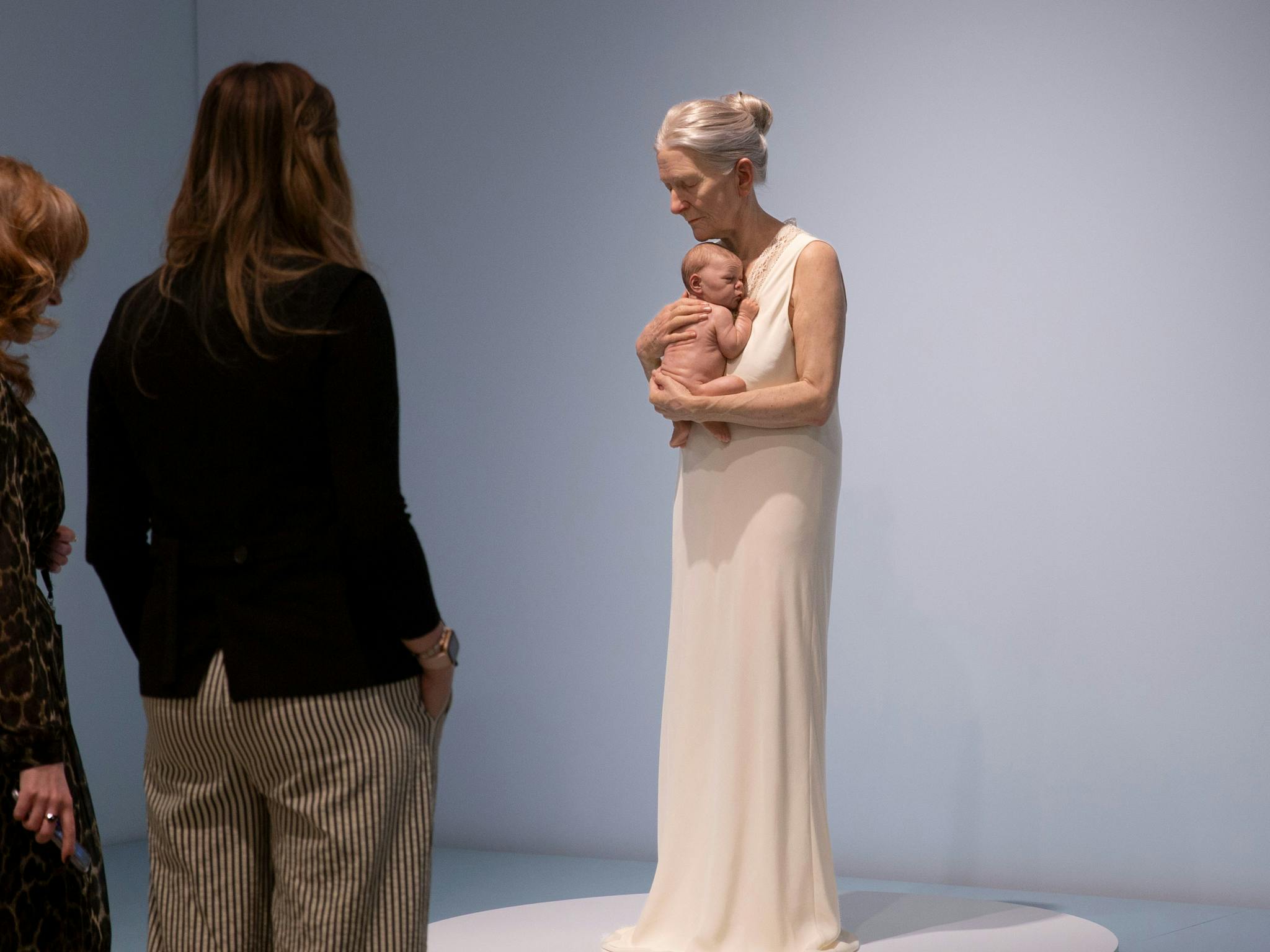 Two women view a hyperrealistic sculpture of an elderly woman in a nightgown holding a baby