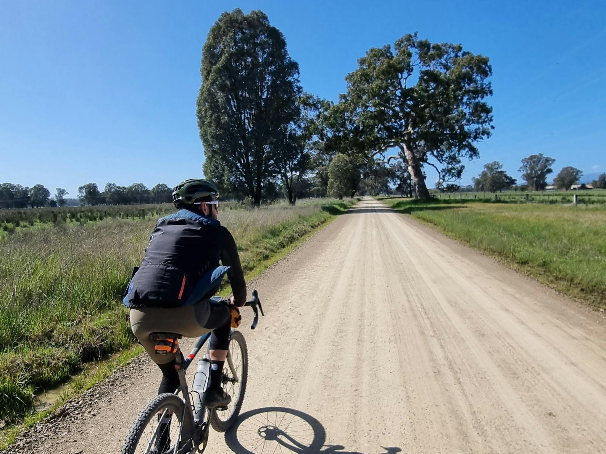Cyclist in foreground on dull brown gravel road, grass, gum trees, blue sky
