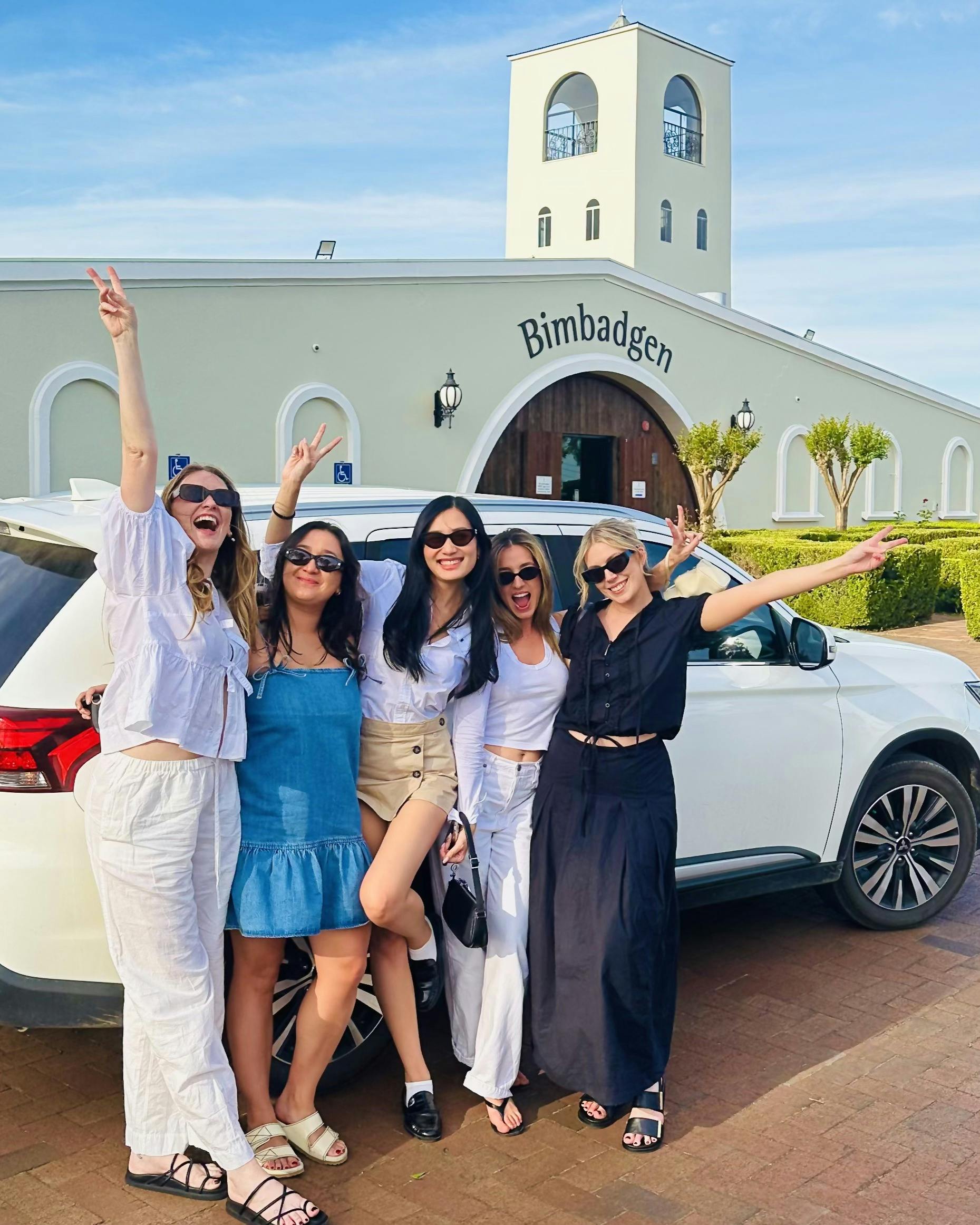 A small group of six smiling tourists standing in front of a modern white luxury transport vehicle o