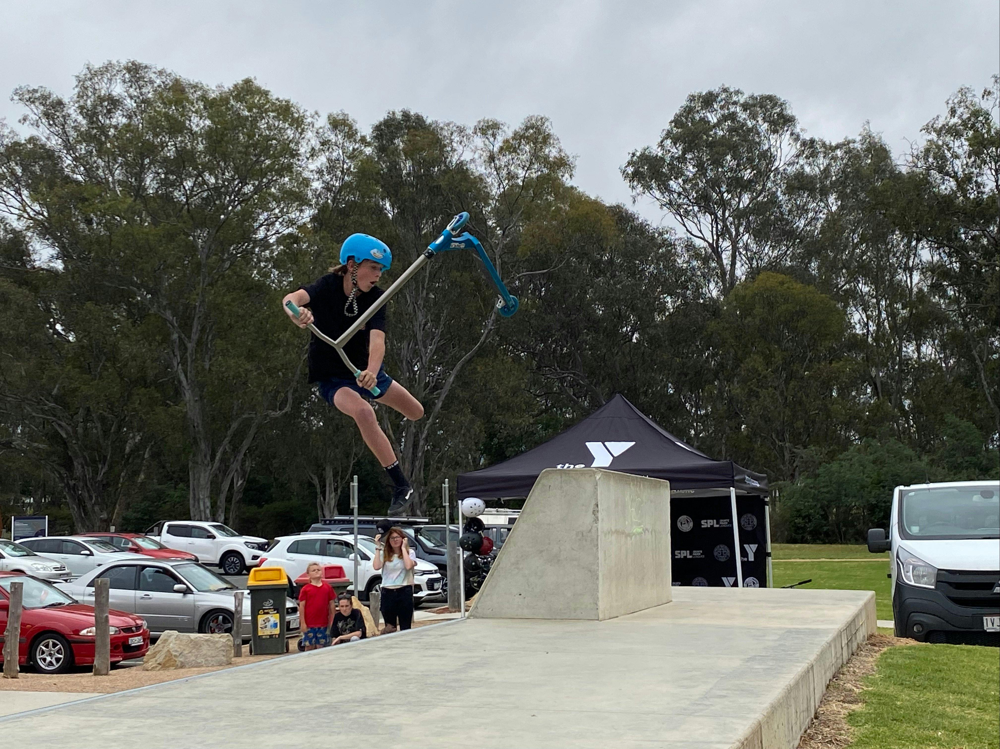 Scooter rider in the air doing a trick above cement people watching cars in background