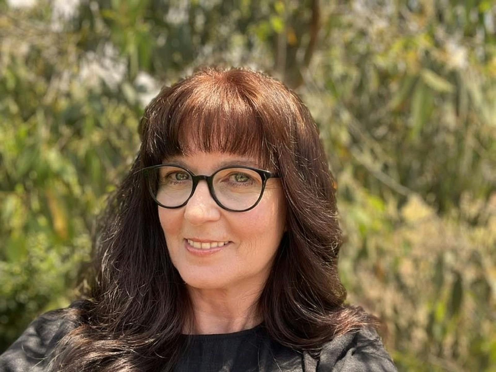 Brunette woman head shot with medium length hair, wearing glasses in front of trees