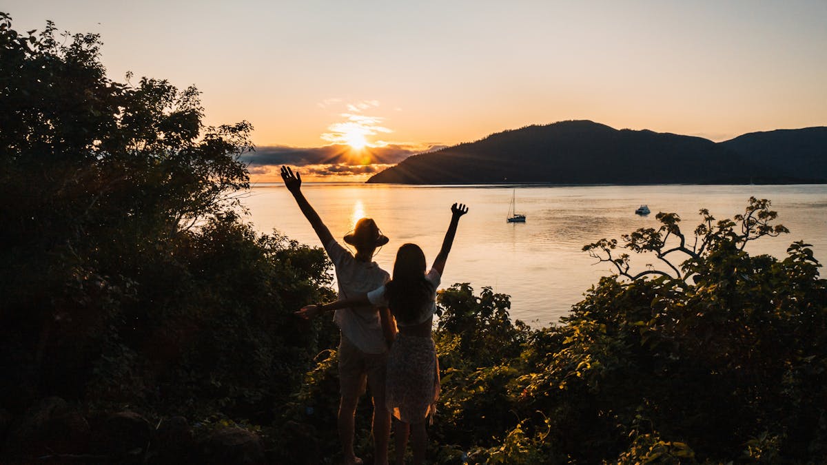 A couple holding hands up on top of a hill facing sunset with an island in the background A couple holding hands up on top of a hill facing sunset with an island in the background