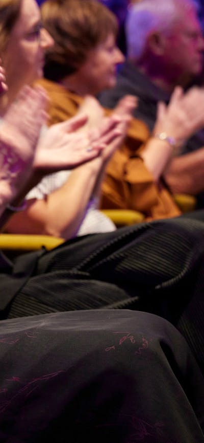 An audience clapping their hands while seated in yellow chairs.