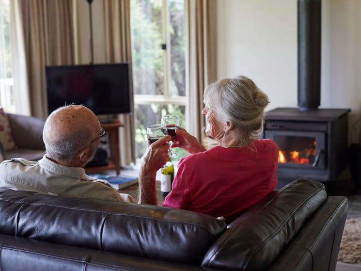 A couple enjoy a glass of wine on a sofa in front of a wood fire inside a cottage
