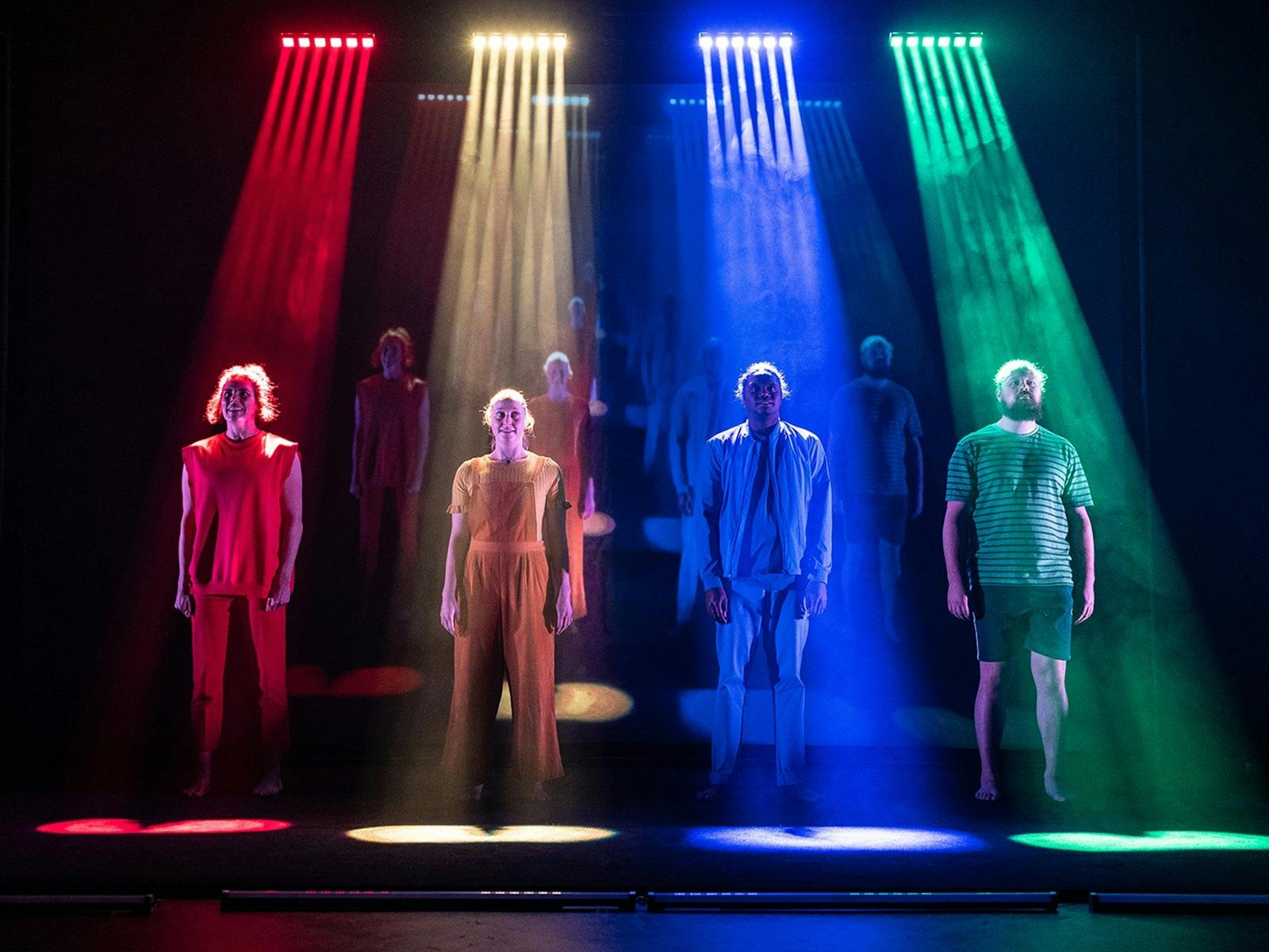 A group of performers stand on a stage, bring coloured lights spotlight each of them