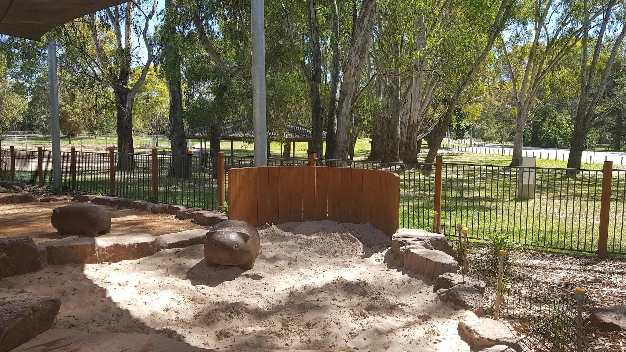 sun shades, childrens playground, wombat sculpture in sandpit, leafy trees, sunshine, fence, rotunda