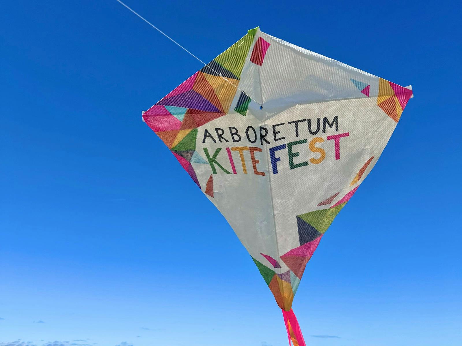 A hand decorated white kite floats in a bright blue sky with the words 'Arboretum Kitefest'