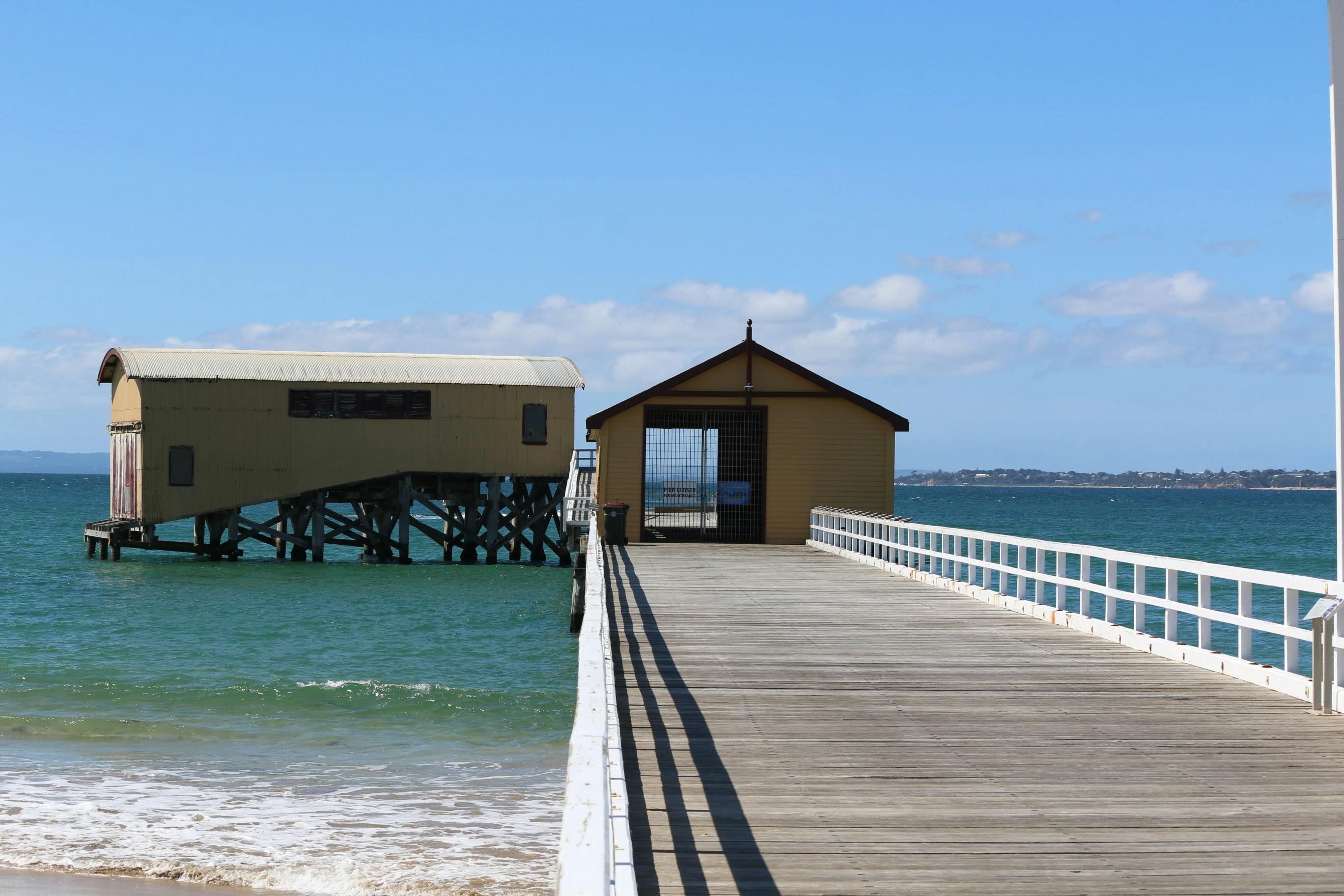 Looking along a timber pier at yellow buildings at the end of the pier. It is sunny.