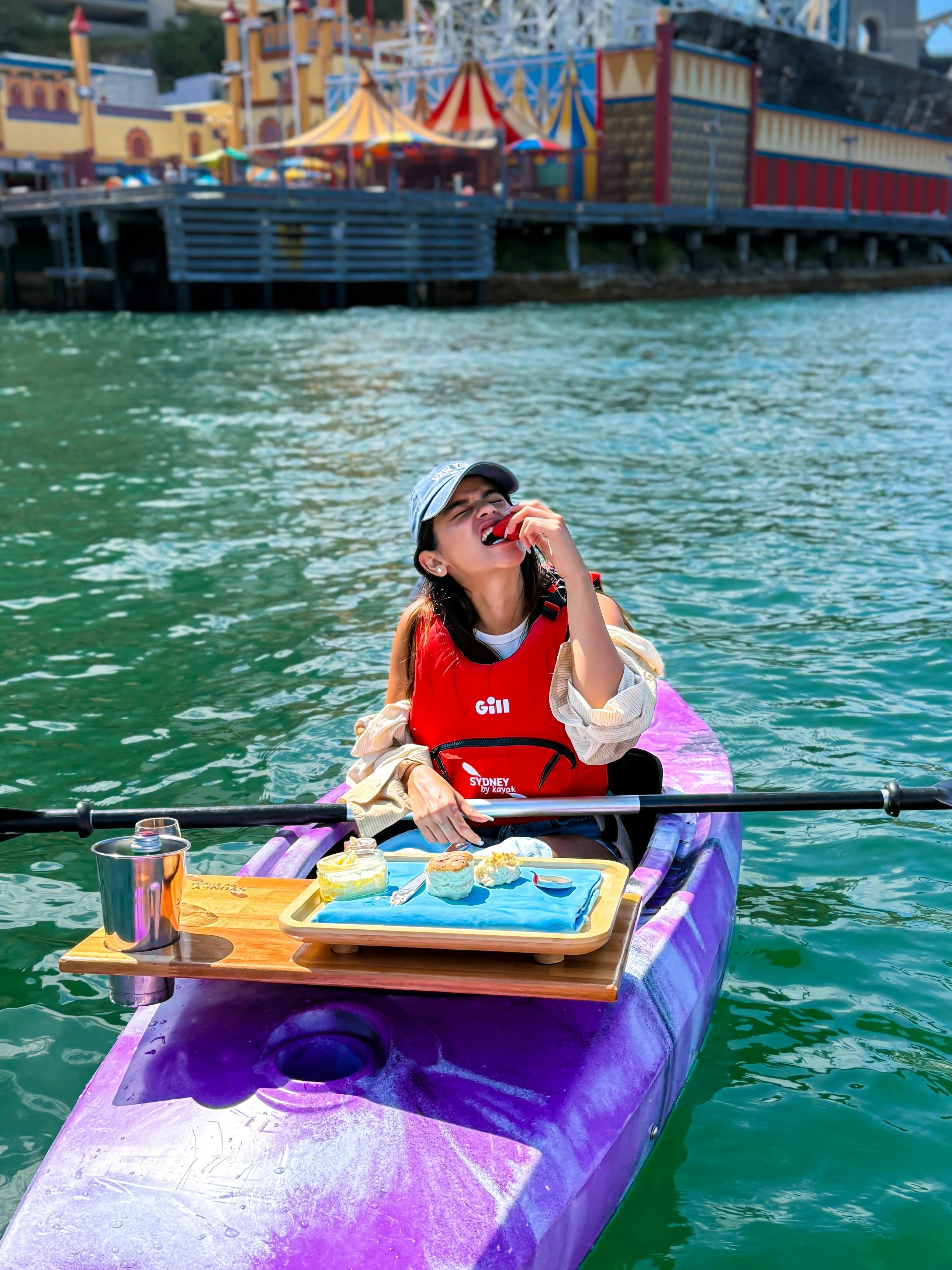 Lady mid bite on a strawberry while sitting on her pink kayak on sydney harbour in the sun.