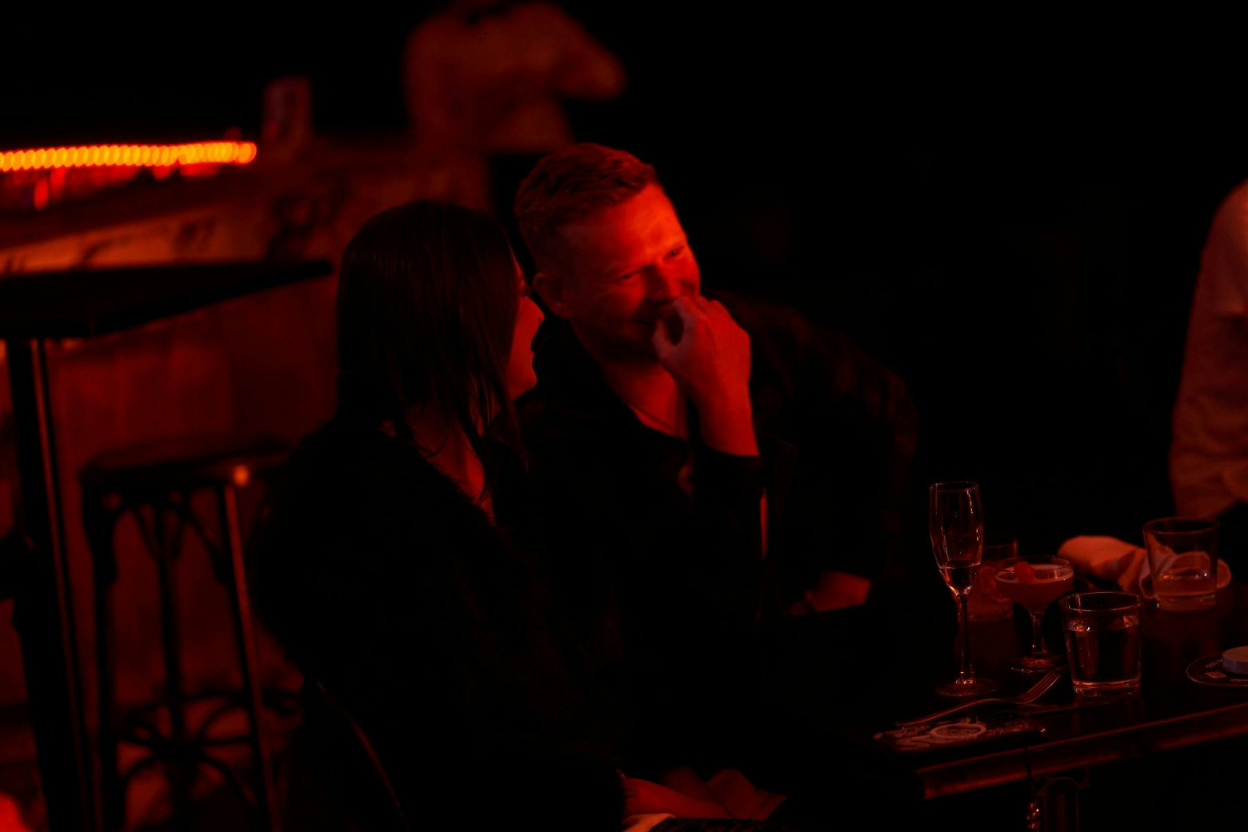 A couple sit in the shadown at their table, leaning close to each other and laughing.