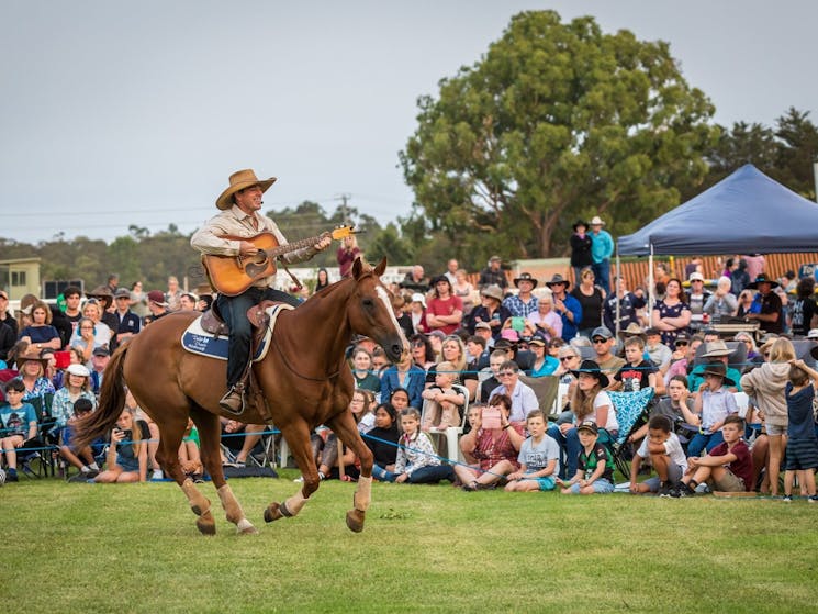 Tom is riding a horse while singing and playing to a crowd of people at an outback show.