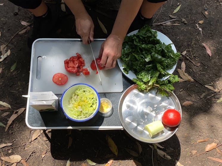 Hands chopping tomatoes on a chopping board with onions and greens adjacent