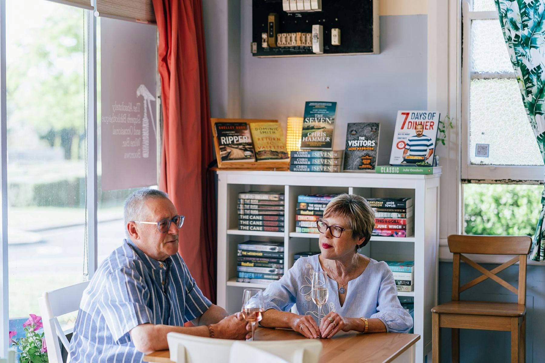 A couple sitting by the window with a glass of wine each. Behind them, a bookshelf of novels.